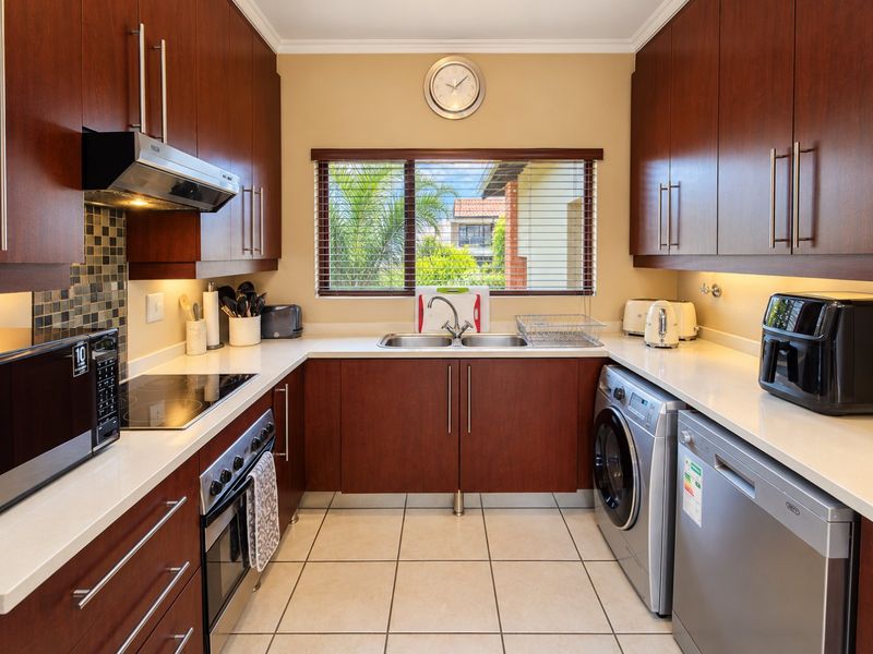 A well-maintained galley kitchen featuring dark wood cabinetry, stone countertops, integrated appliances, and a mosaic tile backsplash with ample under-cabinet lighting.