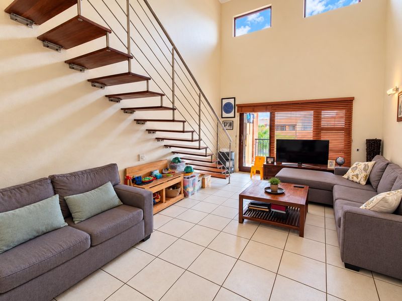 Spacious double-volume living room featuring a modern floating staircase with stainless steel balustrades, tiled flooring, and large windows providing ample natural light.