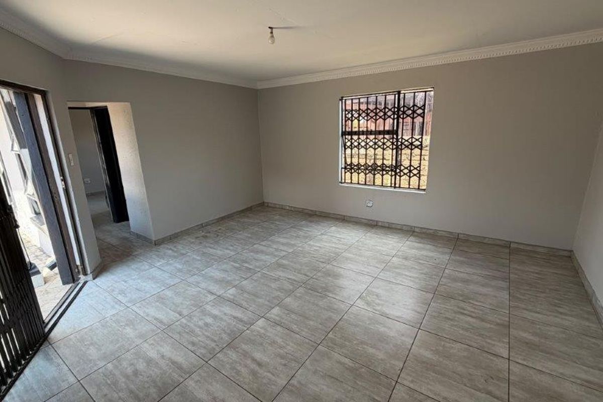 A spacious, empty living area featuring neutral wall tones, tiled flooring, and secure access via a sliding door with a security gate.