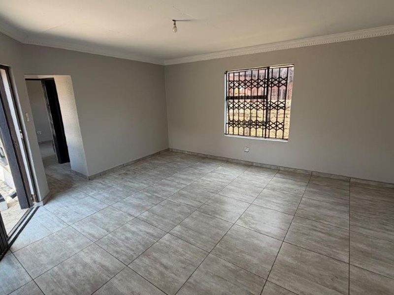 A spacious, empty living area featuring neutral wall tones, tiled flooring, and secure access via a sliding door with a security gate.