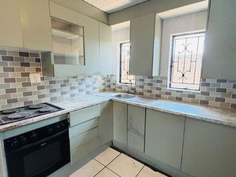 A compact kitchen featuring built-in cabinetry, a tiled backsplash, an integrated oven and hob, and stainless steel sink with security-barred windows.