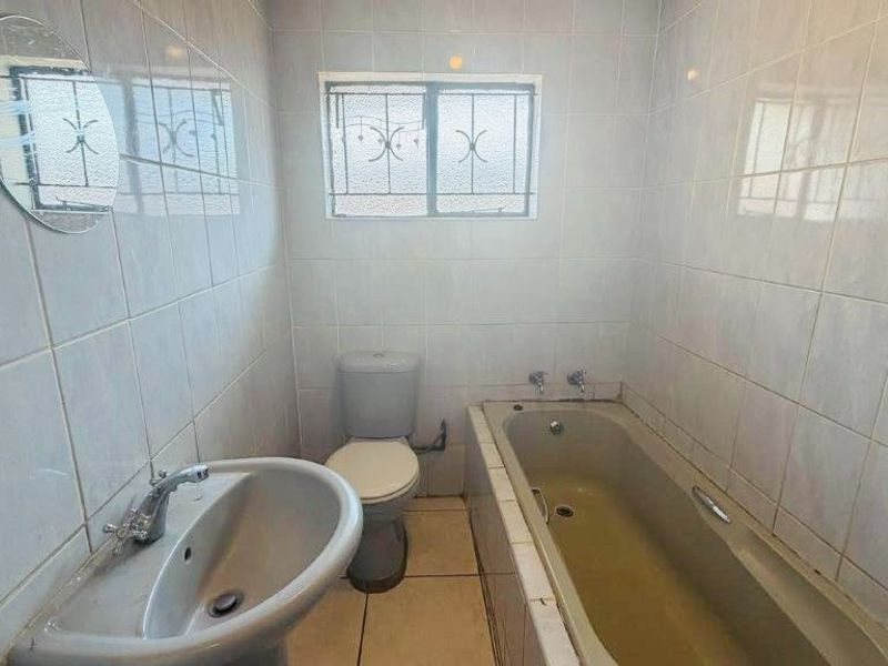 A functional bathroom featuring a pedestal basin, toilet, and bathtub with full-height wall tiling and security bars on the window.