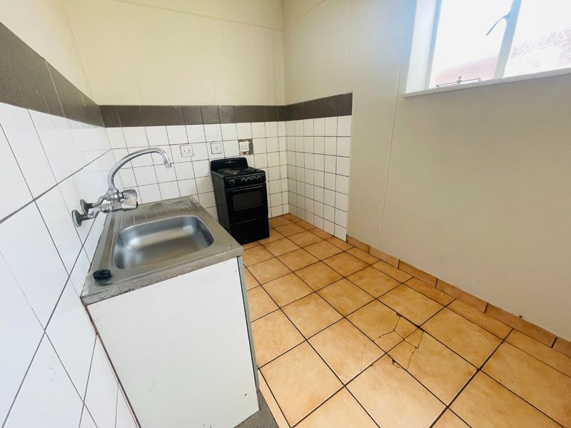 A compact, empty kitchen featuring tiled flooring, a stainless steel sink, and a freestanding stove, requiring some maintenance to the floor tiles.