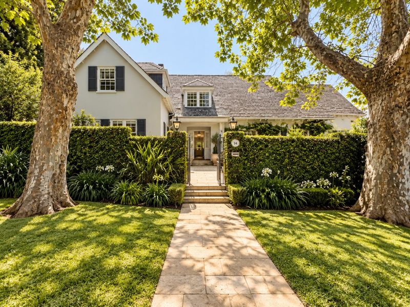 A charming front facade featuring a stone walkway, manicured hedges, and a classic shingle roof, framed by mature trees.