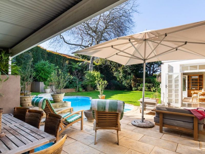 A spacious covered patio overlooking a private swimming pool and lush landscaped garden, featuring paved flooring and direct access to the home via french doors.