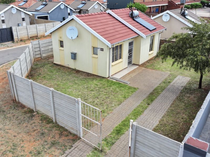 Aerial view of a detached house featuring a tiled roof with solar water heating, a paved driveway, and a fenced garden area.
