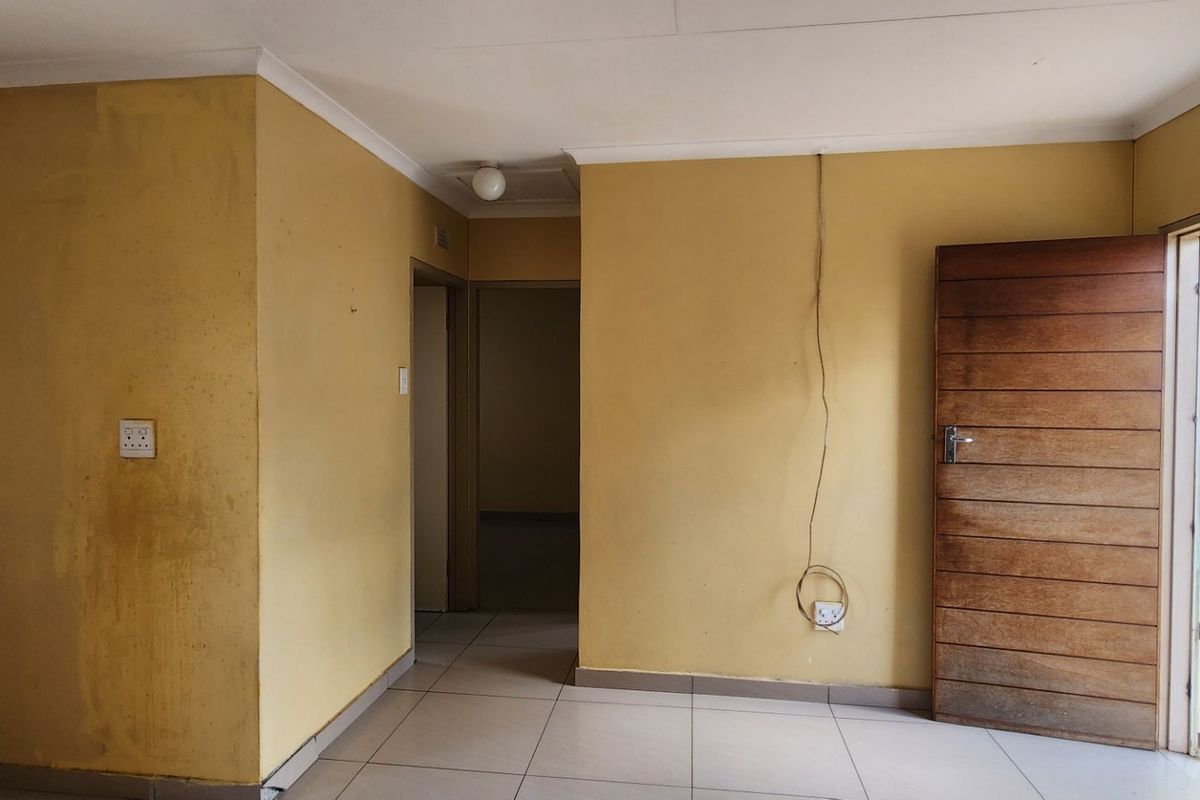 An empty living area featuring tiled flooring, coved ceilings, and a timber door with an external security gate, requiring cosmetic updates.