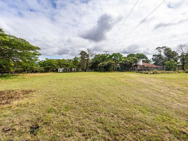A wide-angle view of a large, open grassy field on a farm property, featuring mature trees, perimeter fencing, and a water tank in the distance.