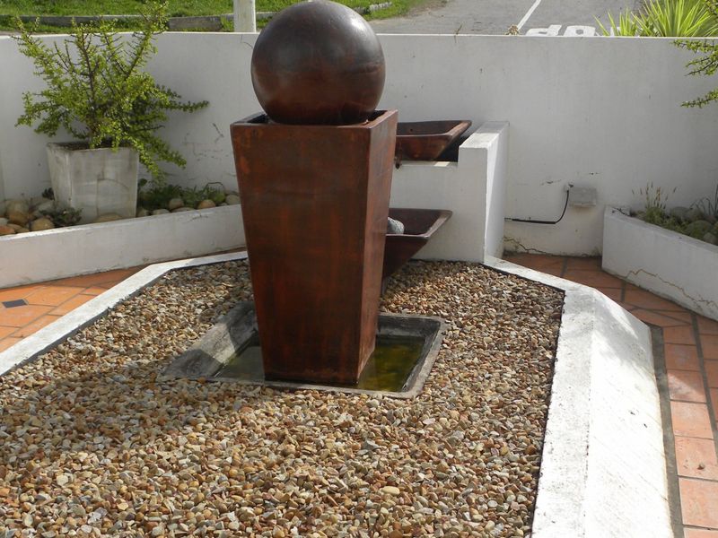 A landscaped garden area featuring a central spherical water feature, gravel beds, and tiled patio surfaces with low retaining walls.