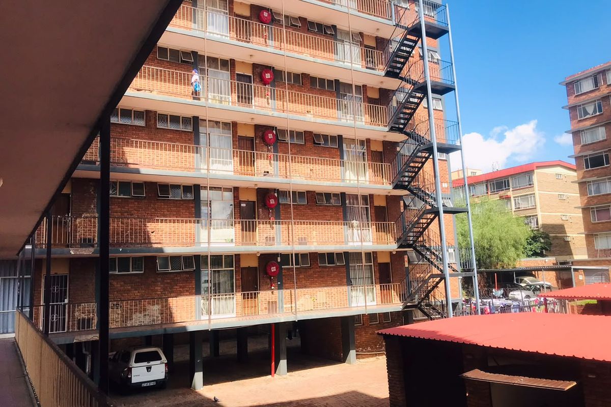 Exterior view of a multi-story apartment building featuring exposed brickwork, external steel fire escape stairs, and ground-level parking area.