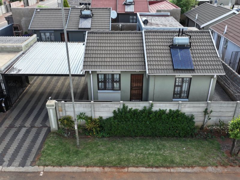 Aerial view of a residential property featuring a tiled roof with solar water heating, a paved driveway, carport, and secure boundary walling.