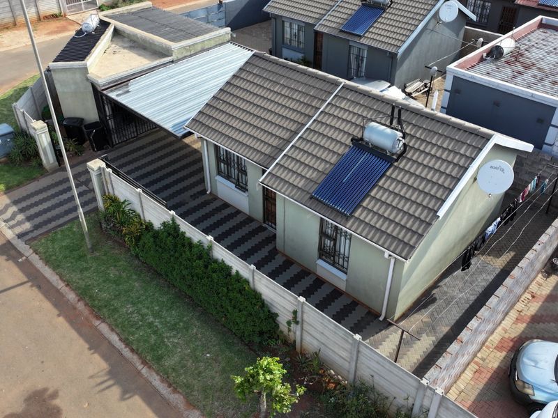 Aerial view of a residential property featuring a tiled roof with a solar water heater, paved driveway, carport, and secure perimeter walling.