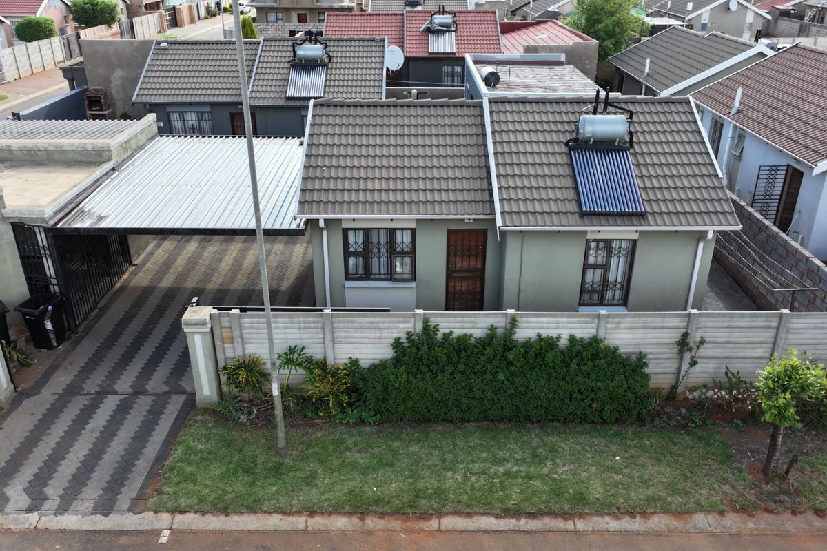 Aerial view of a residential property featuring a tiled roof with a solar water heater, a paved driveway with a carport, and perimeter walling.