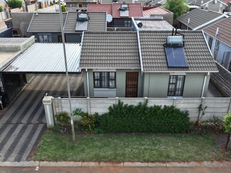 Aerial view of a residential property featuring a tiled roof with a solar water heater, a paved driveway with a carport, and perimeter walling.