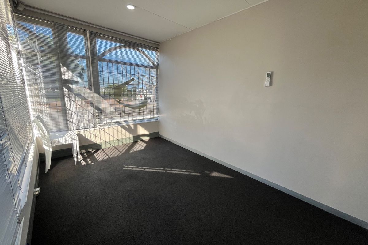 A vacant commercial office space featuring large windows with security bars, carpeted flooring, and a wall-mounted air conditioning control unit.
