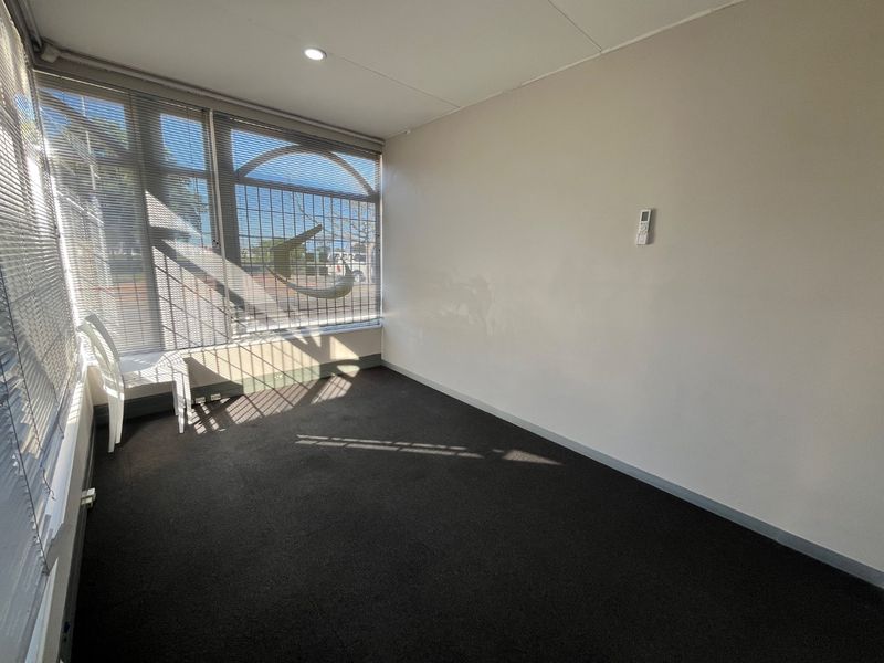 A vacant commercial office space featuring large windows with security bars, carpeted flooring, and a wall-mounted air conditioning control unit.