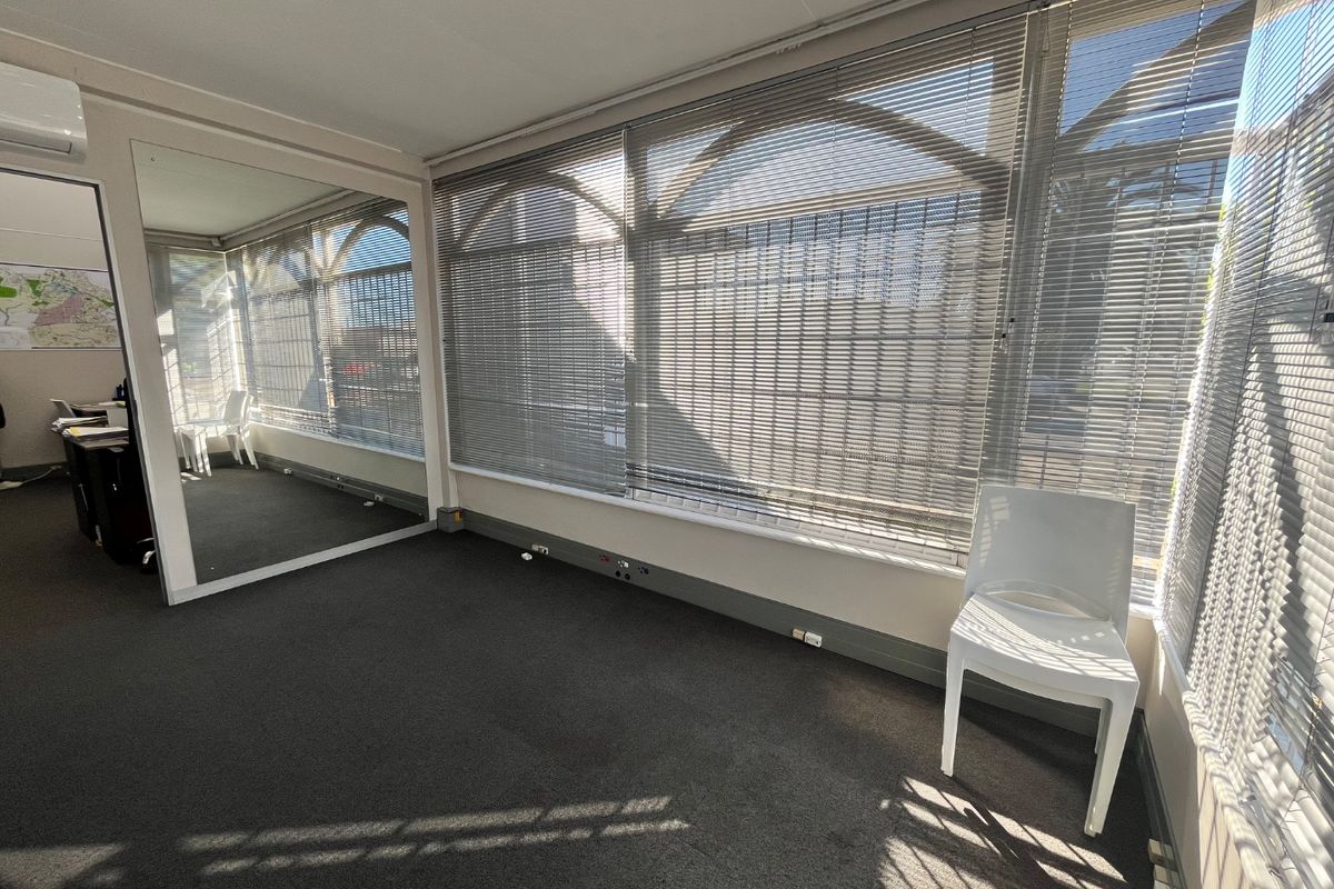 A bright, open-plan office space featuring large windows with venetian blinds, a full-length wall mirror, carpeted flooring, and integrated air conditioning.