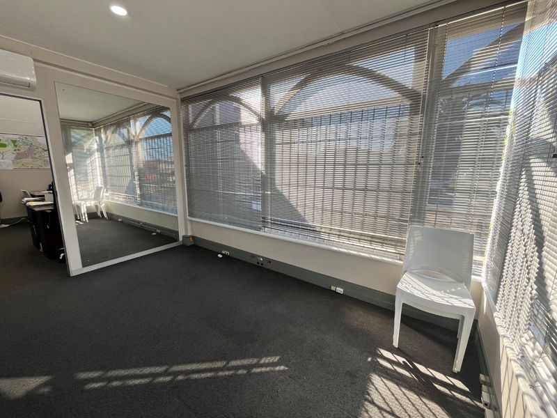 A bright, open-plan office space featuring large windows with venetian blinds, a full-length wall mirror, carpeted flooring, and integrated air conditioning.
