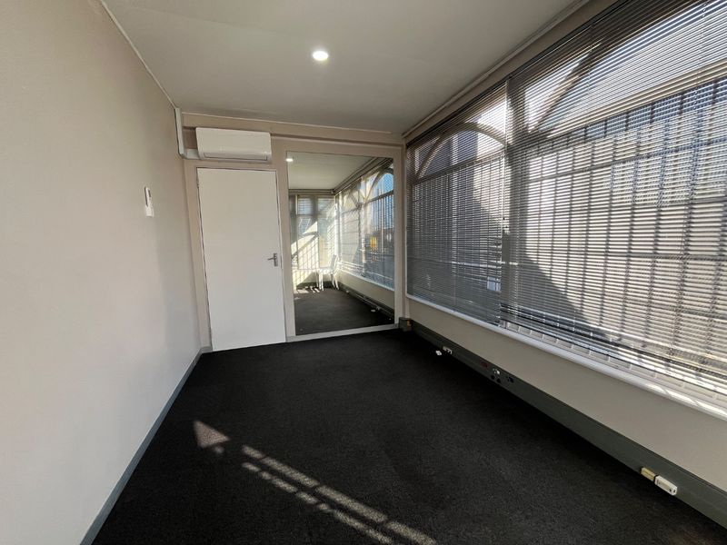 A clean, empty office space featuring wall-to-wall carpeting, a split-system air conditioner, recessed lighting, and large windows with venetian blinds.