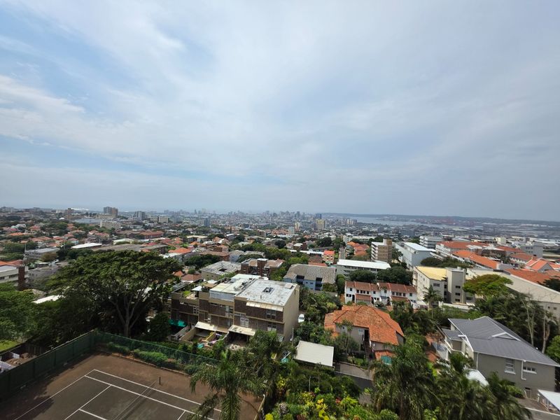Elevated panoramic city and suburban view overlooking residential rooftops and a tennis court.
