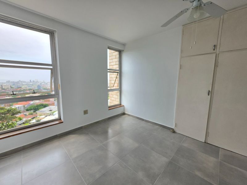 A bright, empty bedroom featuring modern grey floor tiles, built-in cupboards, a ceiling fan, and large windows offering an elevated city view.
