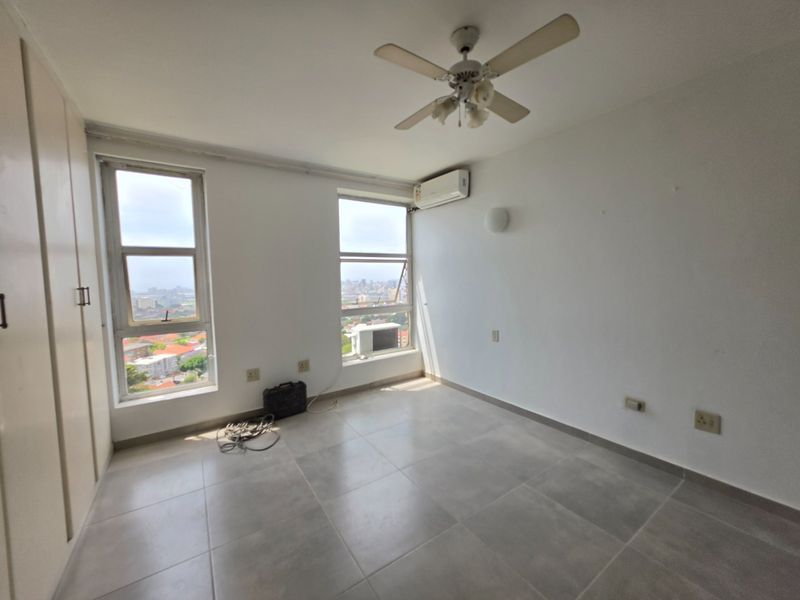 A bright, empty bedroom featuring tiled floors, built-in cupboards, a ceiling fan, and a split-system air conditioner with expansive city views.