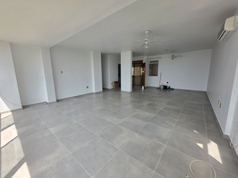 Spacious open-plan living area featuring neutral tiled flooring, ceiling fans, and a wall-mounted air conditioning unit in a clean, vacant setting.