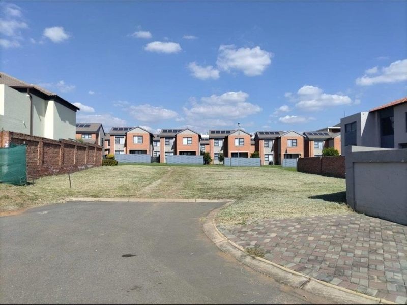 A vacant residential plot featuring a grass lawn, paved driveway access, and brick boundary walls, situated within a developed residential estate.