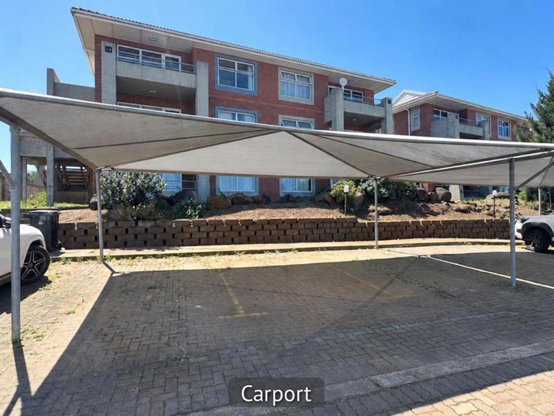 A paved parking area featuring a shade cloth carport structure, situated in front of a multi-story brick apartment building.