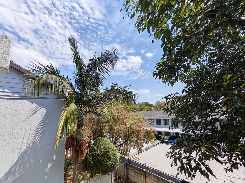 An elevated view overlooking the surrounding residential complex, featuring mature trees, palm foliage, and neighbouring tiled roof structures under a clear sky.