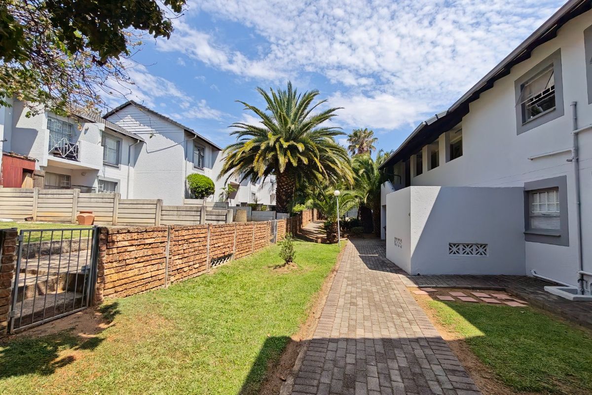 A view of the apartment complex exterior featuring a paved walkway, landscaped lawn, and established palm trees within a secure brick-walled perimeter.