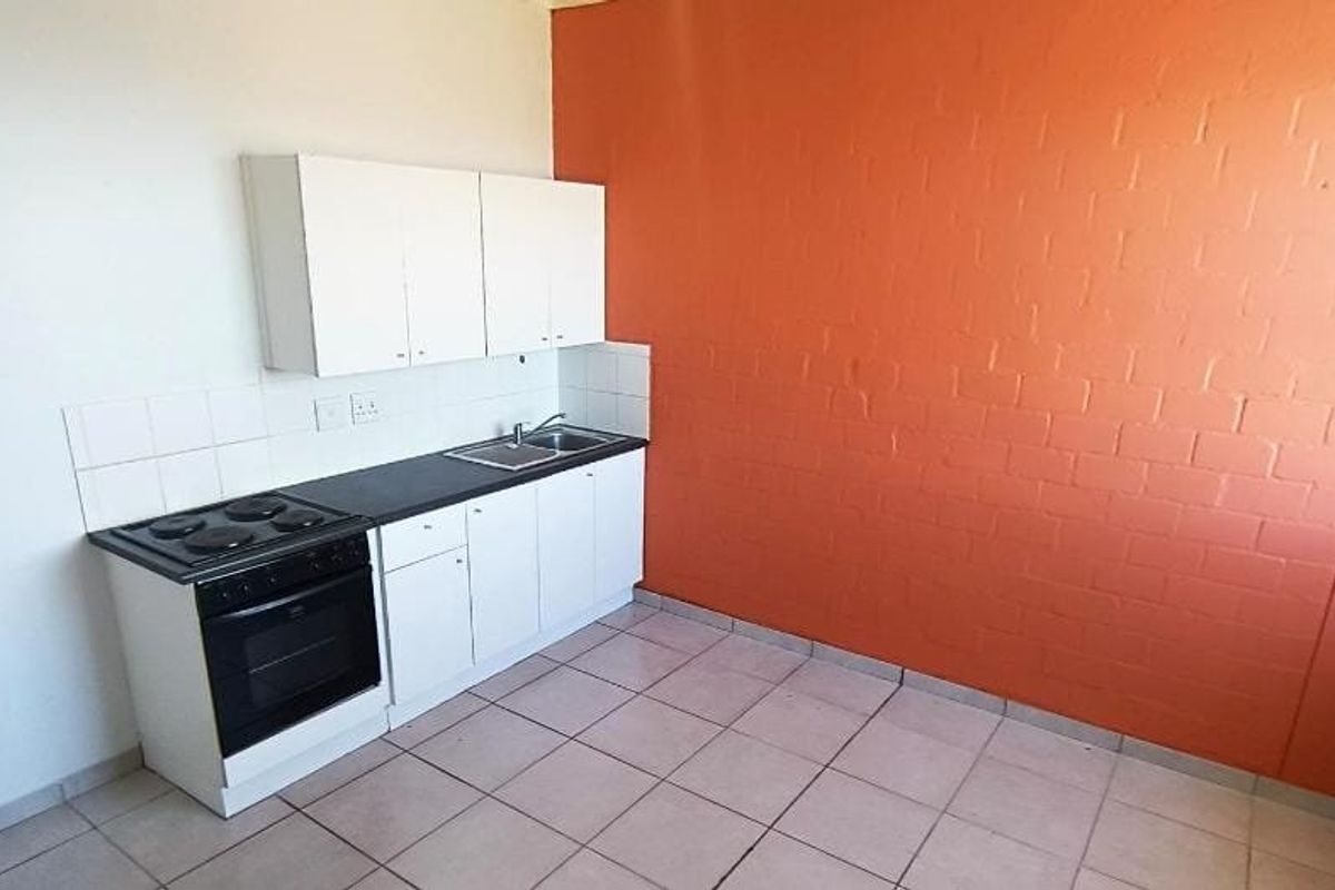 A compact, empty kitchen featuring white cabinetry, a built-in electric stove and oven, stainless steel sink, and tiled flooring with a vibrant orange accent wall.