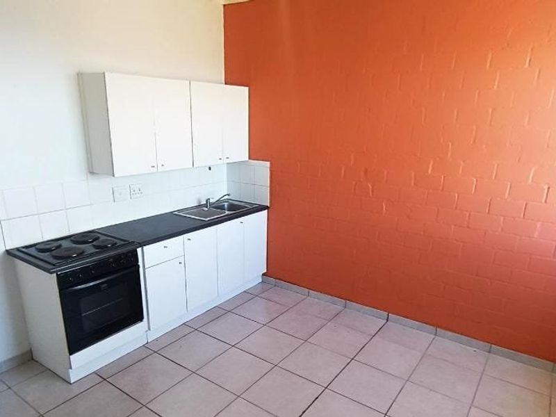 A compact, empty kitchen featuring white cabinetry, a built-in electric stove and oven, stainless steel sink, and tiled flooring with a vibrant orange accent wall.