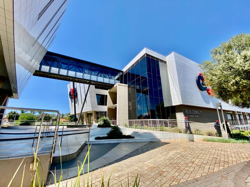 Modern commercial office building exterior featuring a glass curtain wall, elevated pedestrian bridge, and paved entrance area with professional landscaping.