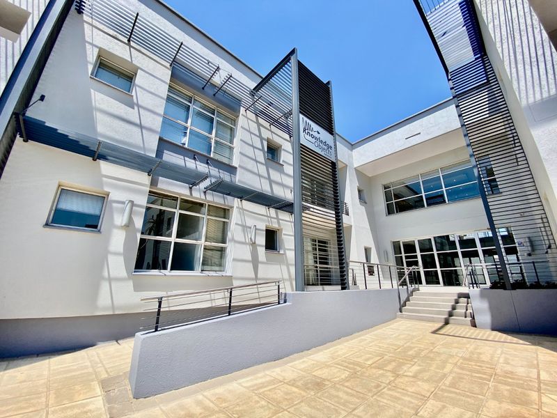 A modern commercial courtyard featuring paved flooring, an accessibility ramp with metal handrails, and architectural metal sunshades over large glass windows.