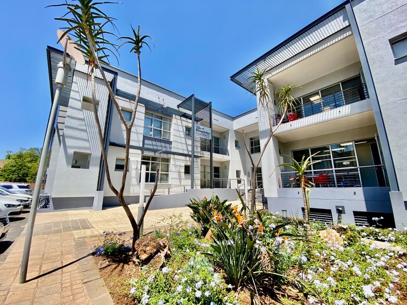 Modern commercial building facade featuring clean grey finishes, large glass windows, balconies with metal railings, and a well-maintained landscaped garden area.