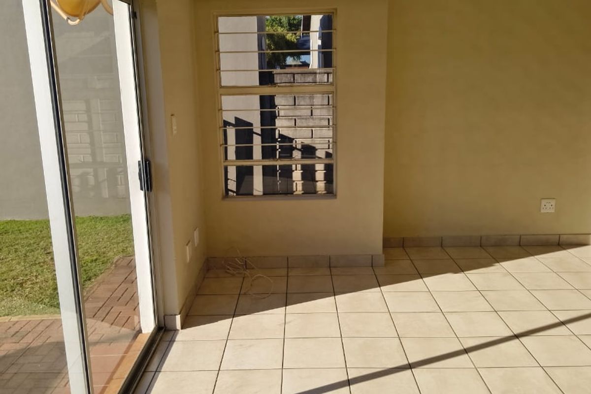 An empty, sunlit living area featuring neutral ceramic floor tiles, a sliding glass door leading to the exterior, and security bars on the window.