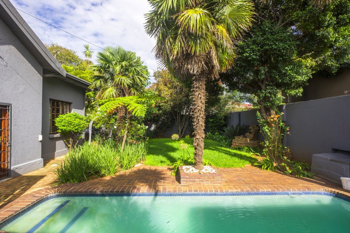 A well-maintained outdoor pool area featuring a swimming pool, brick paved surround, and a lush landscaped garden with mature palm trees.