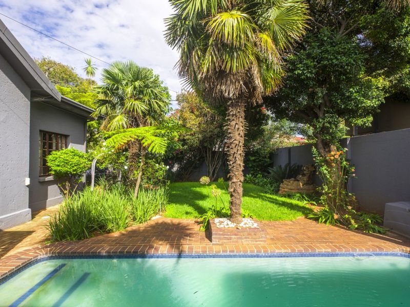A well-maintained outdoor pool area featuring a swimming pool, brick paved surround, and a lush landscaped garden with mature palm trees.