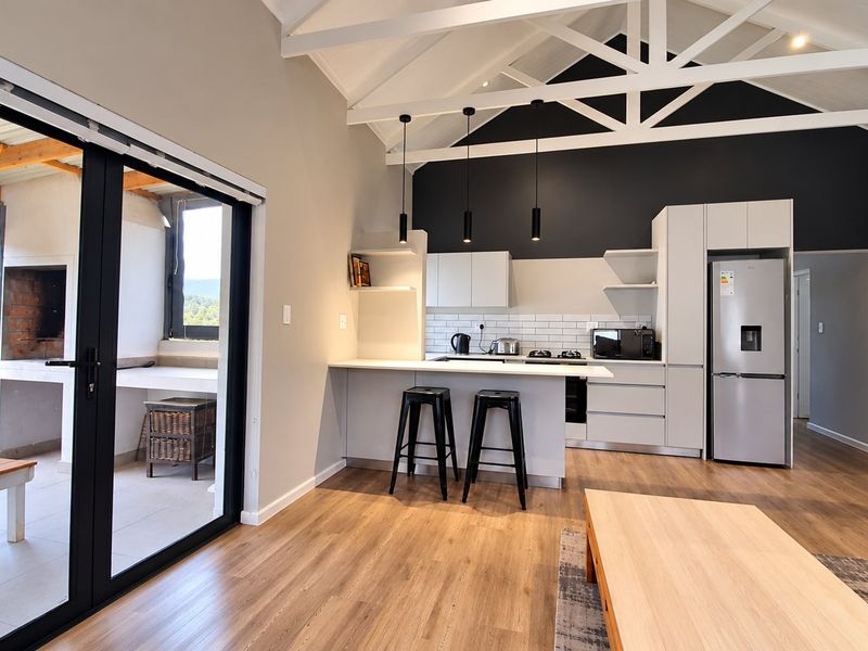 Modern open-plan living area featuring a sleek kitchen with island, vaulted ceilings with exposed trusses, and seamless flow to a covered outdoor patio.