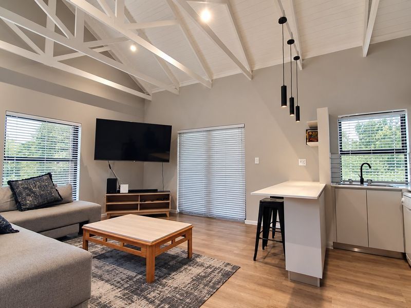 Modern open-plan living area featuring a vaulted ceiling with exposed trusses, integrated kitchen with breakfast bar, and neutral finishes throughout.