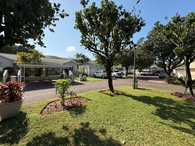 A well-maintained townhouse exterior featuring a paved driveway, carport, and established garden area within a residential complex.