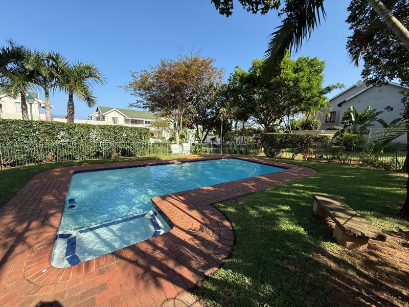 A well-maintained outdoor swimming pool area featuring brick coping, surrounding lawn, and established garden landscaping within a secure residential complex.