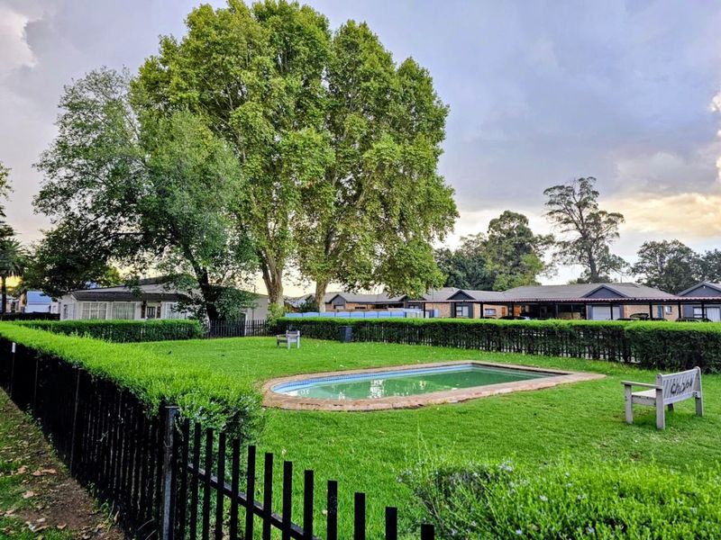 A communal outdoor pool area featuring a swimming pool surrounded by manicured lawns, established trees, and secure metal perimeter fencing.