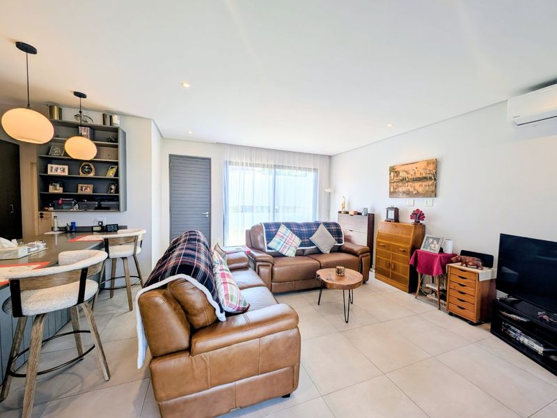 A bright open-plan living area featuring modern tiled flooring, recessed lighting, built-in shelving, and a split-system air conditioner, seamlessly connecting to the kitchen space.