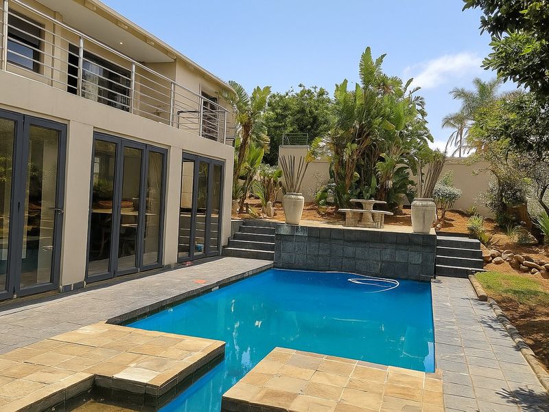 A well-maintained outdoor pool area featuring stone paving, a raised garden terrace with a retaining wall, and direct access to the home via bi-fold doors.