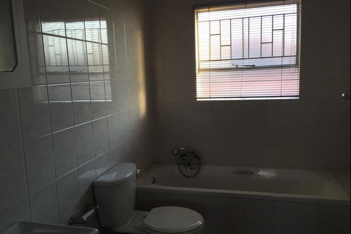 A functional bathroom featuring tiled walls, a standard bathtub, toilet, and basin, with natural light provided by a window fitted with security bars.