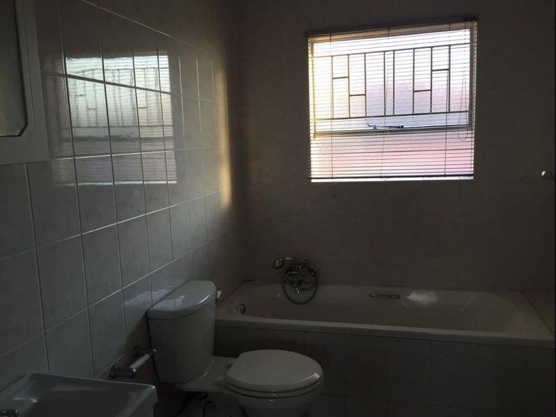A functional bathroom featuring tiled walls, a standard bathtub, toilet, and basin, with natural light provided by a window fitted with security bars.