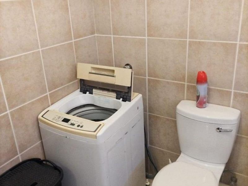A compact laundry area featuring ceramic wall and floor tiling, equipped with a toilet and space for a top-loading washing machine.