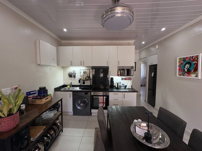 An open-plan kitchen and dining area featuring modern white cabinetry, integrated appliances, tiled flooring, and a contemporary ceiling light fixture.